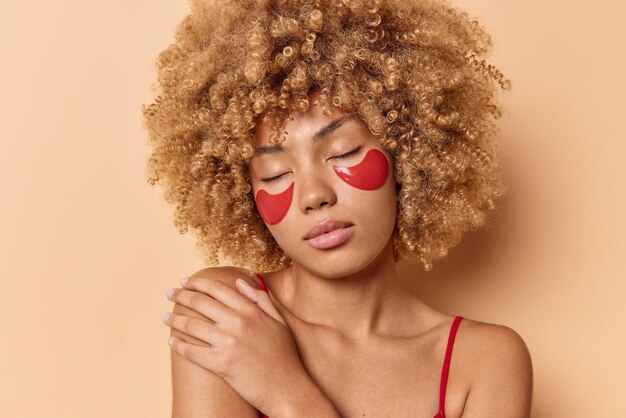 Retrato de encantadora mulher de cabelos encaracolados aplica manchas vermelhas de colágeno sob os olhos para cuidados com a pele e remoção de olheiras toca o ombro suavemente posa contra o fundo bege. Tratamento da zona dos olhos