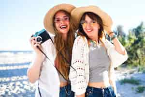 Foto grátis retrato de estilo de vida de verão de meninas de irmã melhores amigos felizes posando na praia, cores claras de sol, chapéus de palha e óculos escuros, segurando a câmera fotográfica vintage, se divertindo juntos.