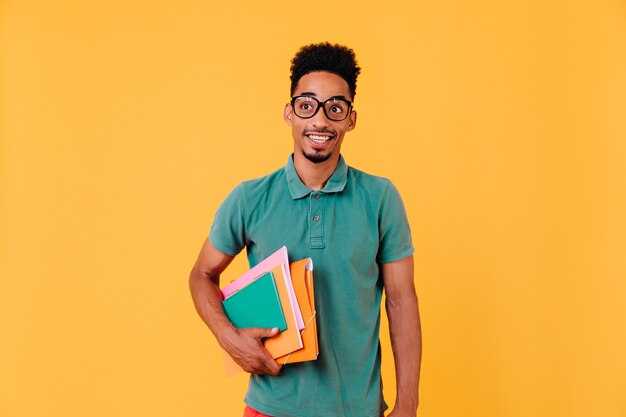 Retrato de estudante africano engraçado em camiseta verde. Foto de feliz menino negro de óculos segurando livros e livros didáticos após os exames.