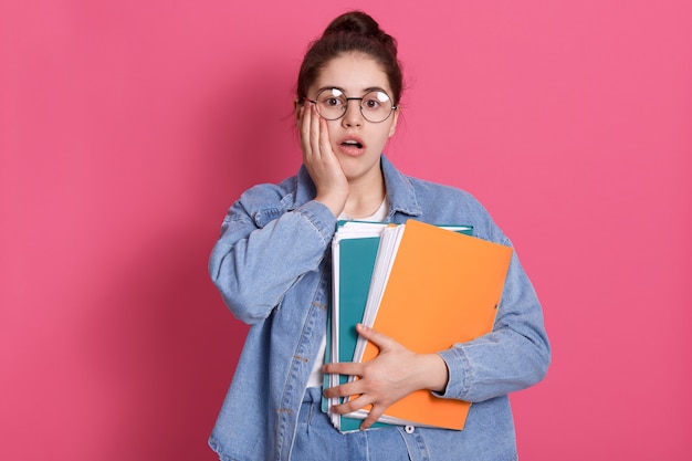 Retrato de estudante com coque de cabelo, usa jeans e óculos arredondados, segurando pastas de papel colorido