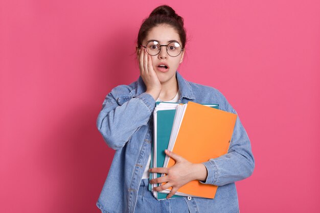 Retrato de estudante com coque de cabelo, usa jeans e óculos arredondados, segurando pastas de papel colorido