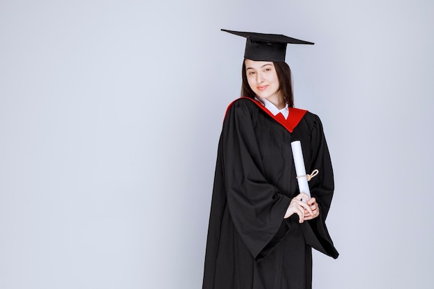 Retrato de estudante de pós-graduação em vestido segurando diploma e em pé. Foto de alta qualidade