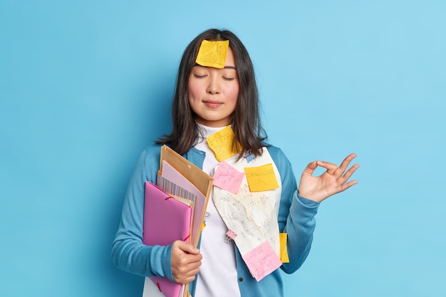 Retrato de estudante relaxado tenta relaxar medita dentro de casa faz gesto de bom, mantém os olhos fechados segura pastas com papéis