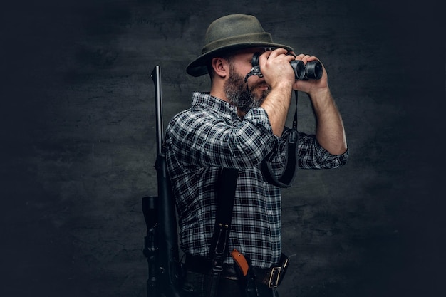 Foto grátis retrato de estúdio de caçador barbudo, vestindo uma camisa de lã xadrez, olhando através de binóculos, segura um rifle.