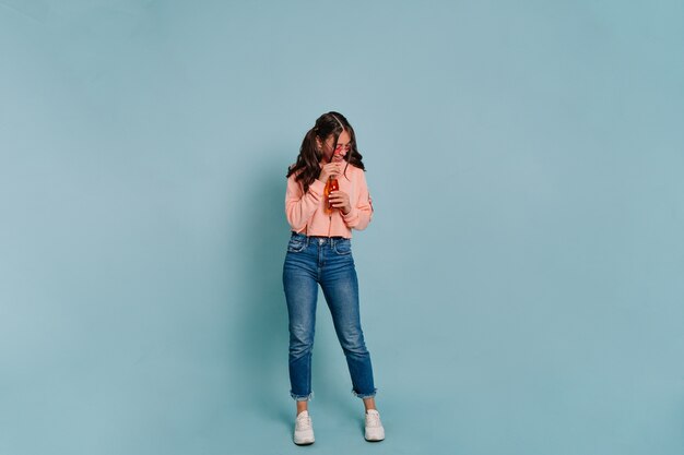 Retrato de estúdio de corpo inteiro de uma mulher moderna e elegante usando pulôver rosa e jeans em óculos rosa, bebendo suco e posando durante a sessão de fotos