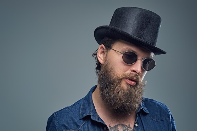 Retrato de estúdio de homem barbudo elegante com uma tatuagem no peito, vestido com uma camisa jeans, chapéu de cilindro e óculos de sol isolados em fundo cinza.