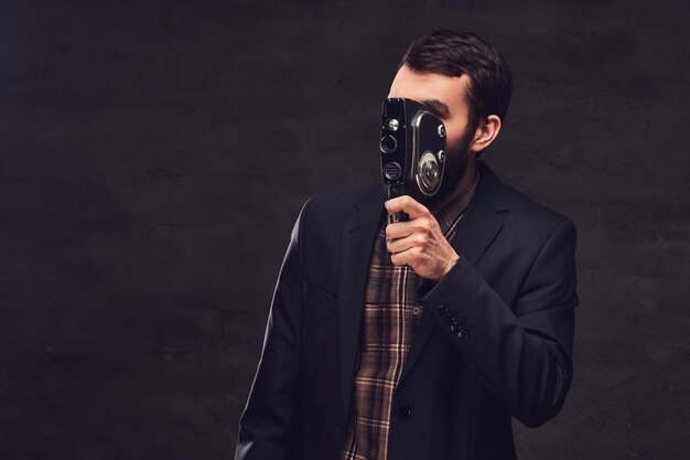 Retrato de estúdio de homem barbudo vestindo um terno clássico segurando uma câmera retro, fazendo uma foto. Isolado em um fundo escuro.