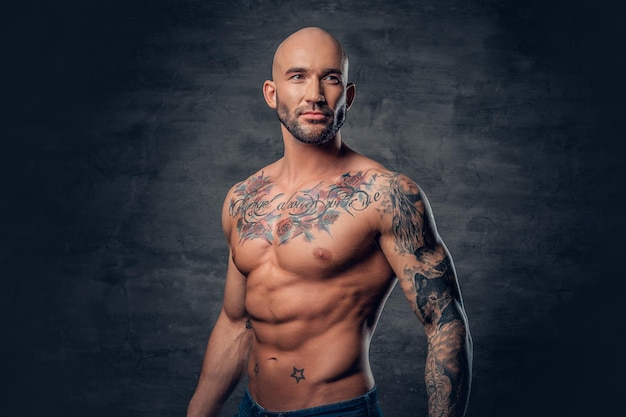 Foto grátis retrato de estúdio de homem de cabeça raspada atlético sem camisa com tatuagens em seu torso posando sobre fundo cinza vinheta.