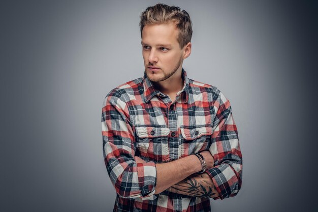 Retrato de estúdio de homem loiro com braços tatuados cruzados, vestido com uma camisa xadrez isolada em fundo cinza vinheta.