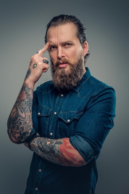 Retrato de estúdio de macho barbudo com tatuagens nos braços, vestido com uma camisa jeans isolada em fundo cinza vinheta.