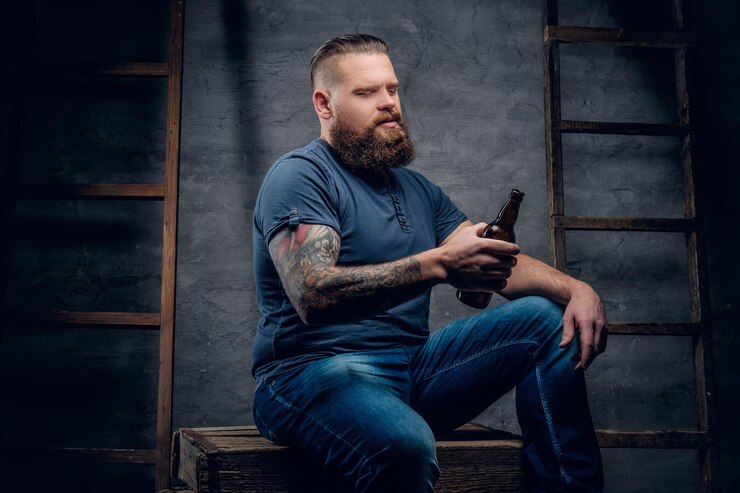 Foto grátis retrato de estúdio de macho hipster tatuado barbudo detém a garrafa de cerveja em fundo cinza com duas escadas de madeira vintage.