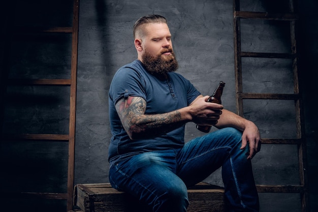 Retrato de estúdio de macho hipster tatuado barbudo detém a garrafa de cerveja em fundo cinza com duas escadas de madeira vintage.