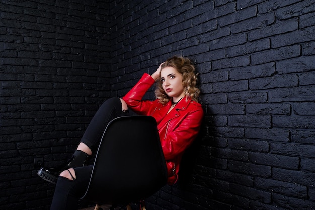 Retrato de estúdio de menina loira na jaqueta de couro vermelha posou na cadeira contra a parede de tijolos