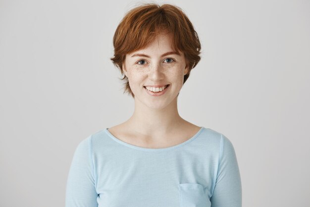 Retrato de estúdio de mulher caucasiana atraente carismática com cor de cabelo ruivo natural e sardas sorrindo alegremente em pé contra um fundo cinza posando para propaganda