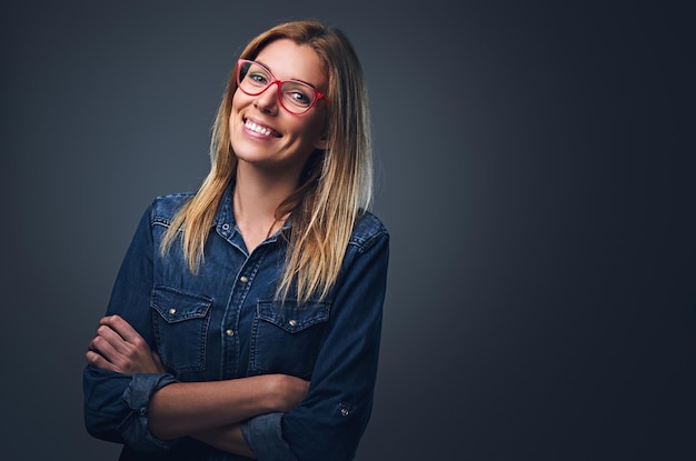 Retrato de estúdio de mulher loira vestida com camisa jeans e óculos vermelhos.