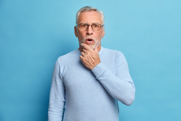 Retrato de estúdio de um homem sênior de cabelos grisalhos chocado segurando o queixo e mantendo a boca aberta ouve algo surpreendente usando um macacão de manga comprida isolado sobre a parede azul