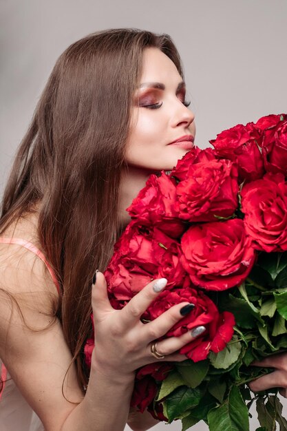 Retrato de estúdio de uma jovem morena deslumbrante com maquiagem usando vestido de noite sexy, cheirando um grande buquê de rosas vermelhas com os olhos fechados, apreciando o cheiro de rosas