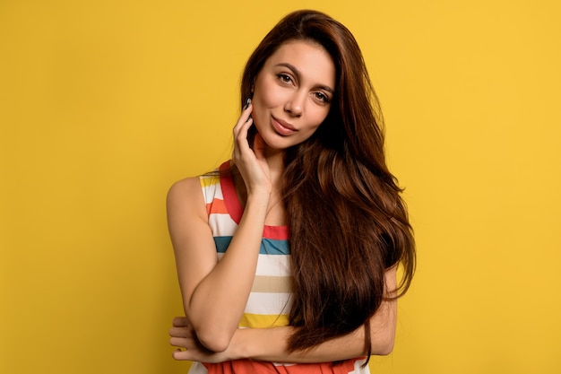 Retrato de estúdio de uma senhora bonita com longos cabelos escuros, usando um vestido de verão brilhante, posando com emoções felizes sobre a parede amarela