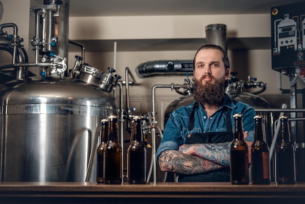 Retrato de fabricante masculino hipster tatuado e barbudo apresentando cerveja na microcervejaria.