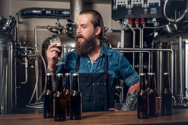 Retrato de fabricante masculino hipster tatuado e barbudo, degustação de cerveja na microcervejaria.