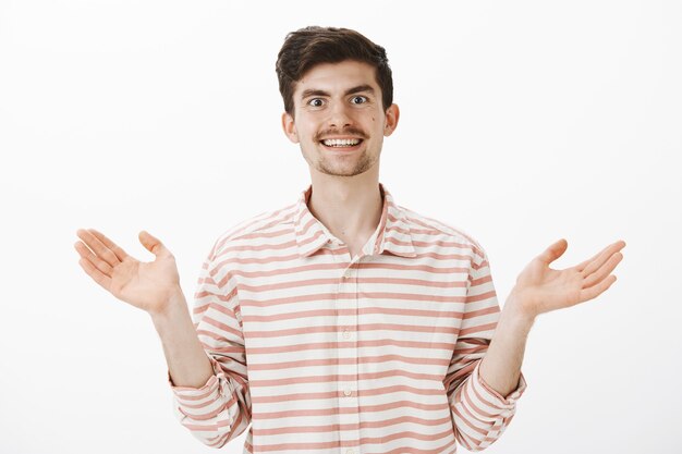 Retrato de feliz despreocupado e atraente colega de trabalho em camisa listrada, levantando as palmas das mãos e sorrindo amplamente, expressando atitude positiva e amigável, não impressionado e indiferente à situação