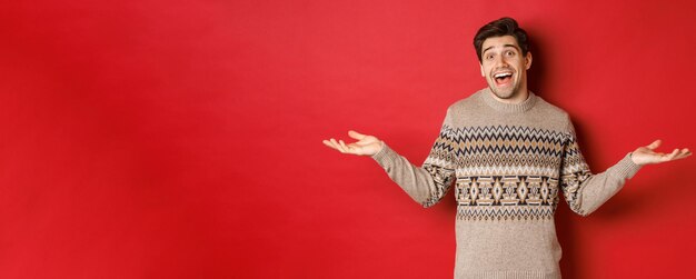 Retrato de feliz e surpreso, cara caucasiano bonito, vestindo suéter de Natal, espalhou as mãos de lado e parecendo sem noção, em pé sobre um fundo vermelho.