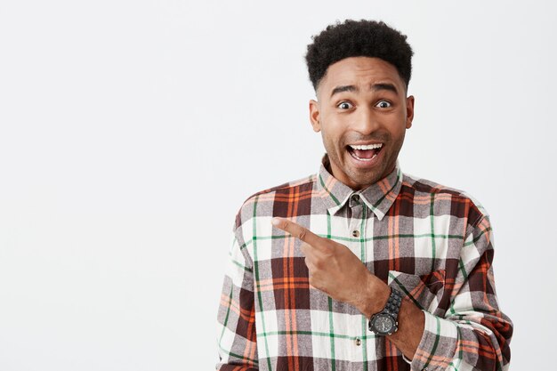 Retrato de feliz jovem estudante masculino de pele bronzeada com penteado afro em camisa quadriculada casual, sorrindo, apontando de lado com o dedo com a expressão do rosto animado