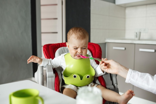 Retrato de filho recém-nascido infeliz no babador verde chorando e se recusando a comer da colher enquanto a mãe pacientemente tenta alimentá-lo.