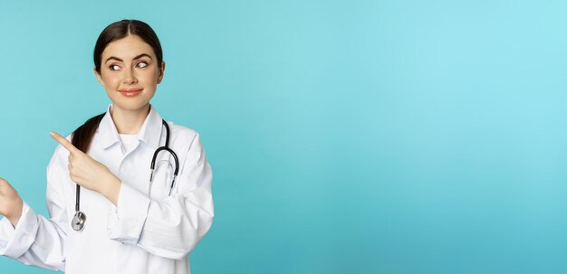 Retrato de funcionária médica sorridente, médica de casaco branco com estetoscópio apontando os dedos para a esquerda mostrando publicidade de clínica médica fundo turquesa