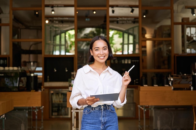 Retrato de gerente de mulher asiática em pé com tablet na frente da entrada do café dá as boas-vindas aos convidados