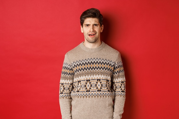 Retrato de homem adulto confuso com suéter de Natal, desapontado durante as férias de ano novo de pé, não entendo algo, em pé sobre fundo vermelho.