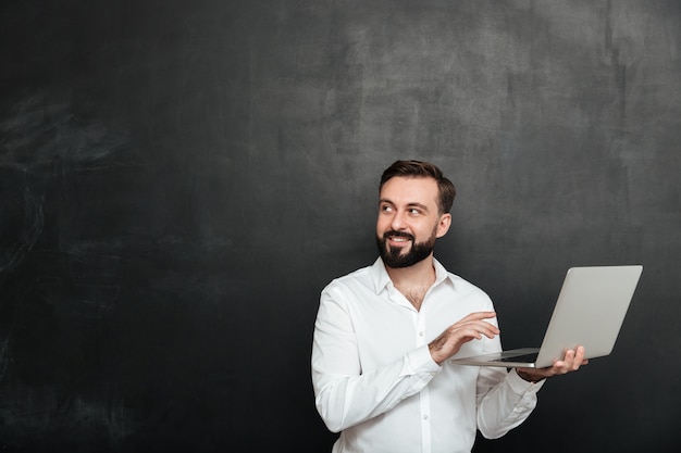Retrato de homem adulto sorridente segurando laptop prata e olhando de lado, isolado sobre a parede cinza escura