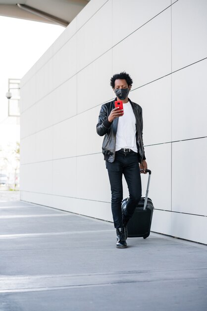 Retrato de homem afro-turista digitando no telefone e carregando a mala enquanto caminha ao ar livre na rua