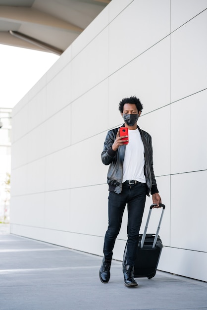 Retrato de homem afro-turista usando seu telefone celular e carregando a mala enquanto caminha ao ar livre