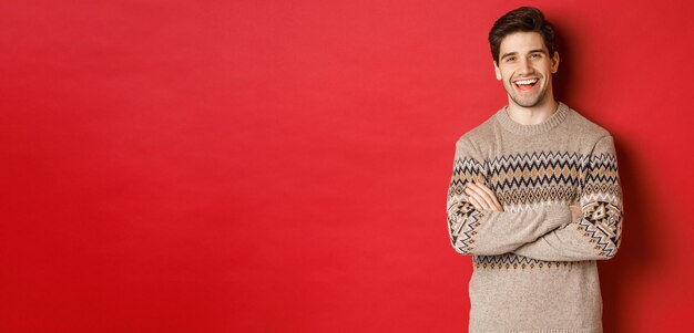 Retrato de homem alegre e atraente na camisola de natal, rindo e sorrindo, comemorando o ano novo e feriados de inverno, de pé sobre fundo vermelho.