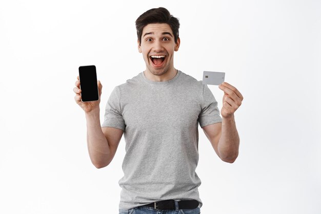 Retrato de homem animado mostrando a tela vazia do telefone e o cartão de crédito demonstram a loja on-line e fazem o pedido em pé sobre fundo branco