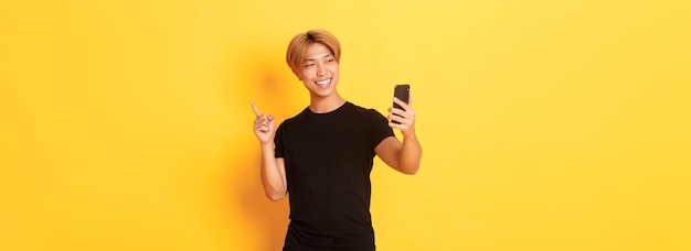 Retrato de homem asiático sorridente bonito tendo chamada de vídeo no smartphone e apontando o dedo para algo