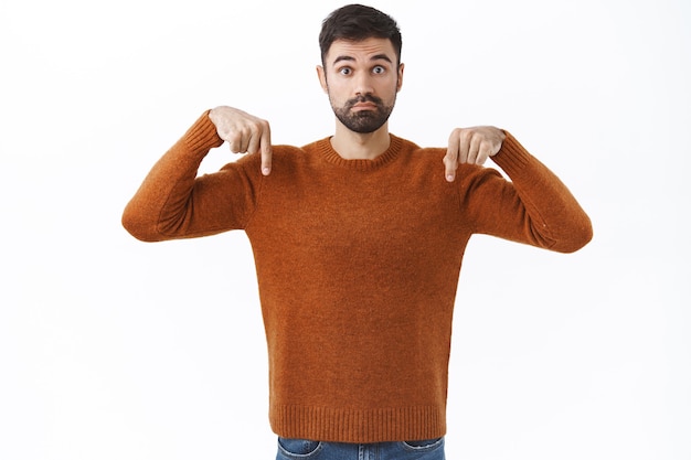 Foto grátis retrato de homem barbudo bonito indeciso fazendo perguntas sobre o produto, apontando o dedo para baixo inseguro, consultando a namorada, pergunte a opinião, parede branca