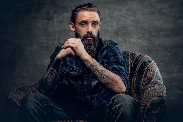 Retrato de homem barbudo com braços tatuados, vestido com uma camisa de flanela xadrez senta-se em uma cadeira sobre fundo cinza escuro.