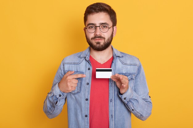 Retrato de homem barbudo confiante apontando com o dedo indicador para cartão de crédito, pagando com cartão para compra