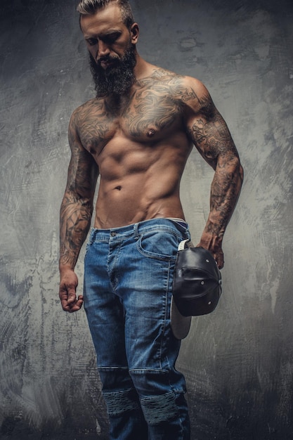 Retrato de homem barbudo tatuado em jeans e torso nu sobre fundo cinza.