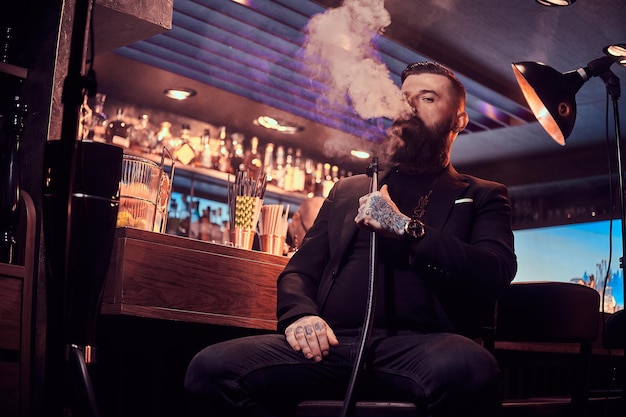 Foto grátis retrato de homem barbudo tatuado que está fumando cachimbo de água, fazendo bom vapor enquanto está sentado perto do balcão do bar.