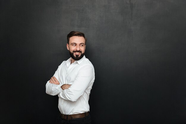 Retrato de homem bonito alegre em pé com os braços cruzados e olhando para trás sobre o espaço da cópia cinza escuro