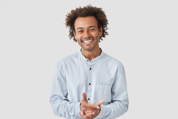 Retrato de homem bonito alegre mantém as mãos juntas, sorri amplamente, vestido com uma camisa elegante