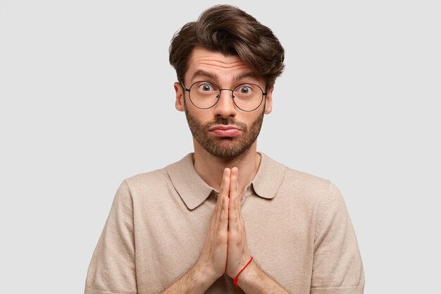 Retrato de homem bonito com aparência europeia, mantém as mãos em gesto de oração, pede perdão com expressão miserável, franze os lábios, vestido casualmente