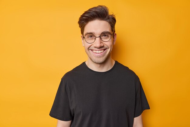 Retrato de homem bonito de cabelos escuros sorri com dentes se sente feliz por usar óculos redondos e camiseta preta casual expressa emoções positivas isoladas sobre fundo amarelo Conceito de felicidade