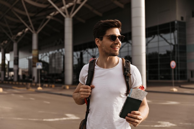 Retrato de homem bonito em camiseta branca e óculos de sol detém passaporte de mochila e bilhetes e poses perto do aeroporto de bom humor