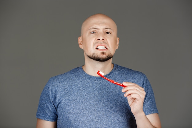 Retrato de homem bonito engraçado na camisa cinza, escovar os dentes ao longo da parede escura