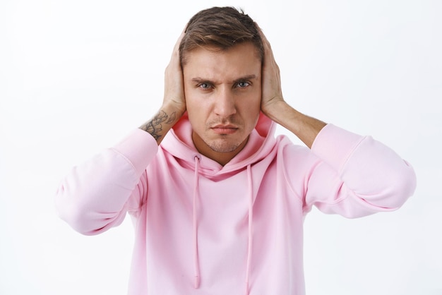 Retrato de homem bonito irritado com raiva fecha os ouvidos e olha a câmera com aversão de desânimo não suporta vizinho barulhento tocando música muito alto em pé irritado fundo branco