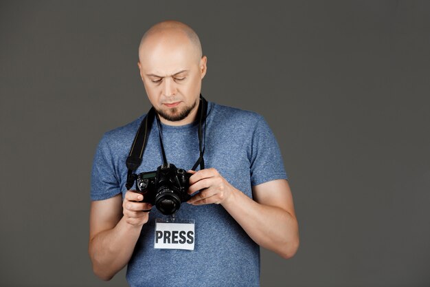 Retrato de homem bonito na camisa cinza com photocamera e imprensa distintivo tirando fotos sobre parede escura